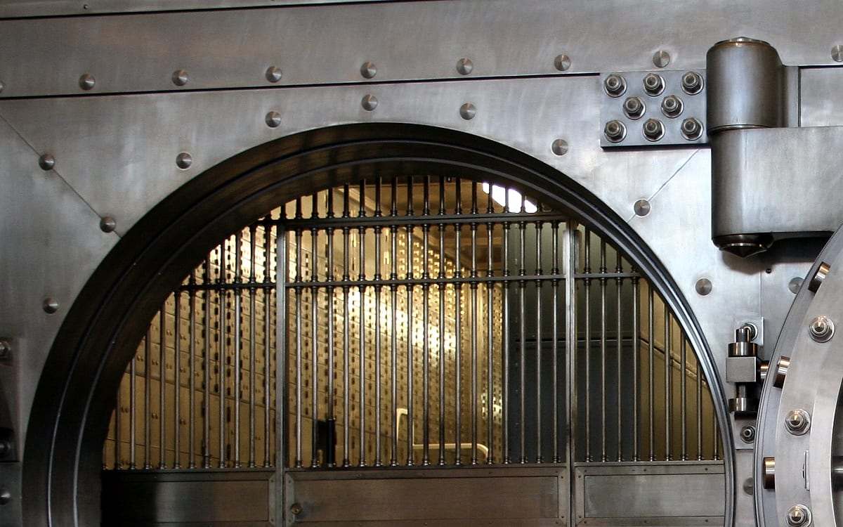 The open arched door of a bank vault, its steel frame studded with rivets, a barred gate drawn across the lit interior behind it.