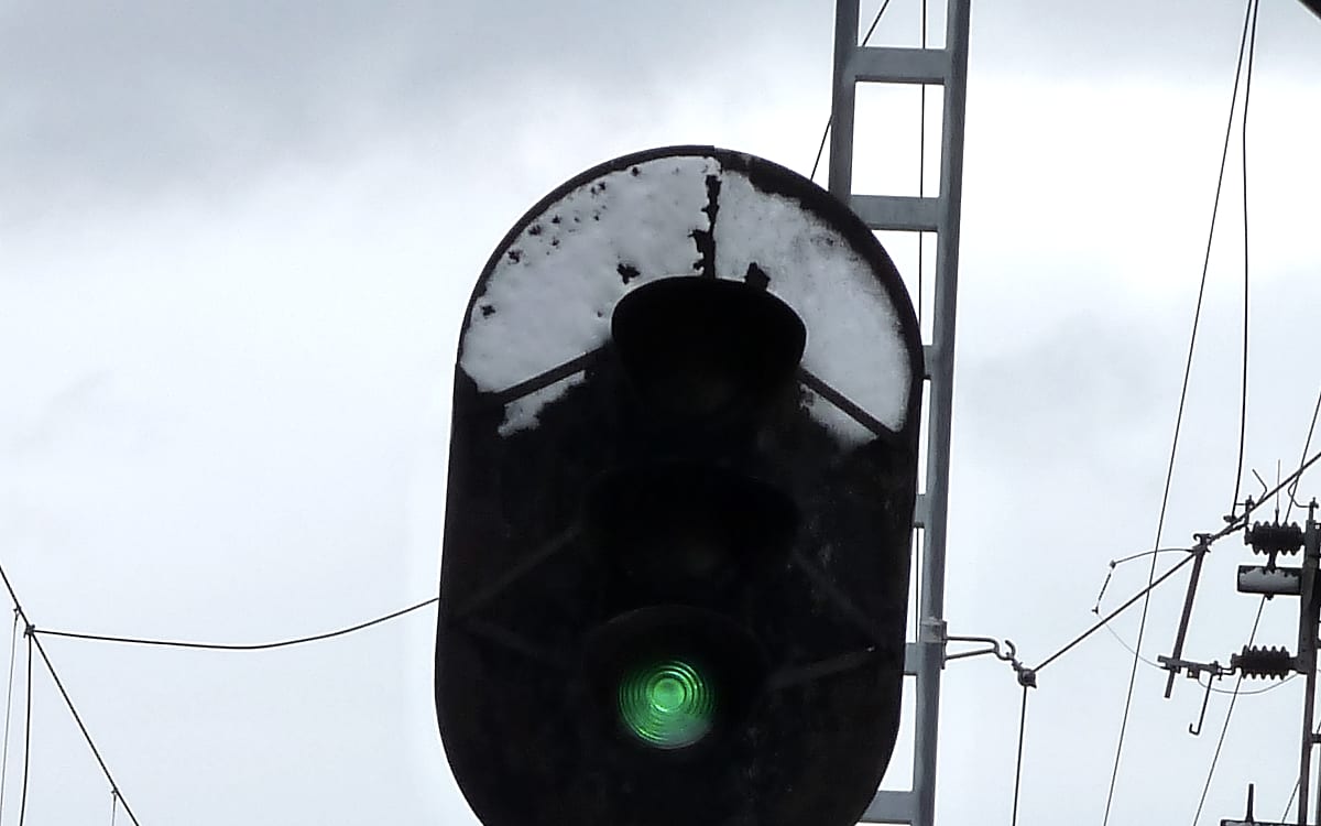 A railway signal head capped with snow against a flat gray sky, its lower lamp lit bright green, overhead wires crossing the frame behind it.